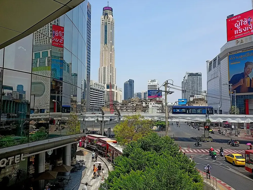 Baiyoke Tower rising above the Pratunam skyline, seen from a raised walkway over a road junction in daylight
